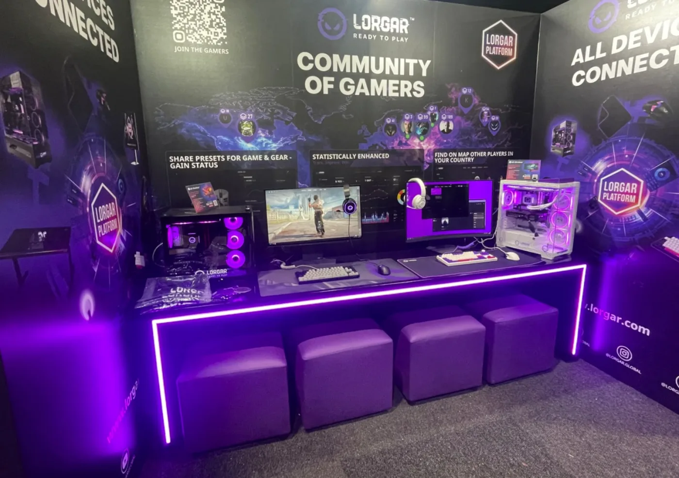 The LORGAR activation pod, lit in purple. Two monitors, keyboards, a headset and two lit PC towers along a counter, with purple seating in front. The back wall reads COMMUNITY OF GAMERS above a world map of players.
