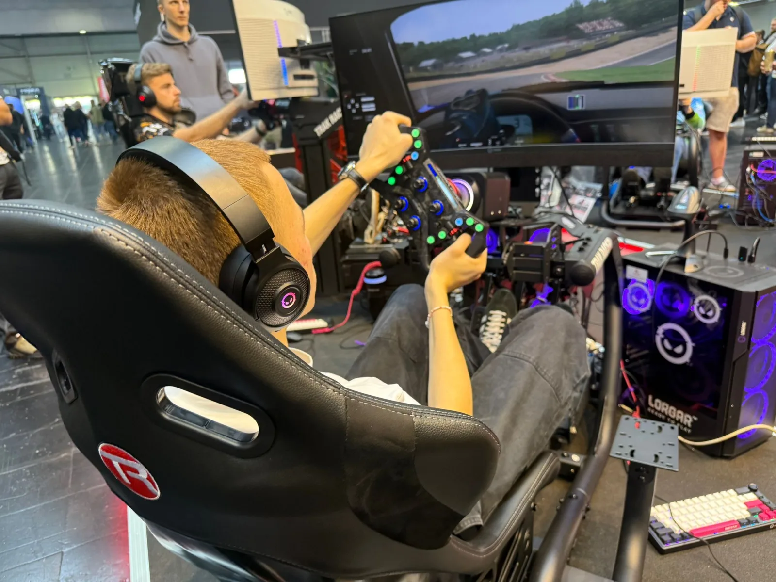 A player in a LORGAR racing rig at a stand, wearing LORGAR headphones, hands on the wheel, racing on the screen in front of them. More rigs and people behind.